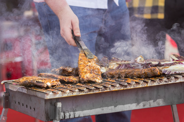 Person grilling ribs and vegetables on an outdoor barbecue with visible smoke.