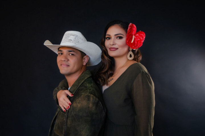 Man in cowboy hat and woman with red flower pose against dark background.